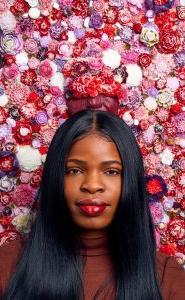 Portrait of a woman with long black hair. Behind her are bright glass flowers, ranging in varying shades of pinks and purples.